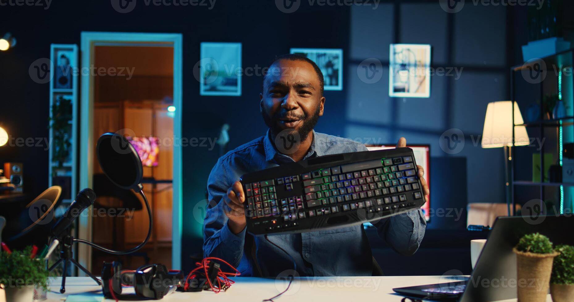 Influencer in dimly lit living room uses camera to film keyboard, mouse and headphones review for online streaming platforms. Online star hosts internet show, unboxing wired gaming peripherals photo