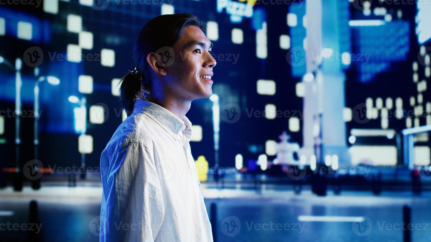 Relaxed asian man wandering around city streets at night, looking upwards to surrounding skyscrapers in metropolitan urban setting. Businessman leaving work, taking a walk outside photo