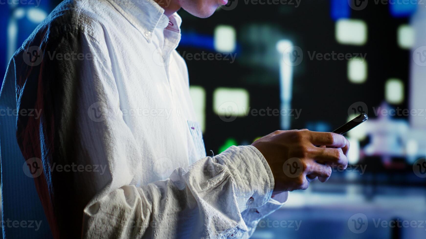 Man walking around city buildings at night, scrolling on social media using phone, blurry cityscape background. Businessman strolling outside in empty intersection, using smartphone, close up photo