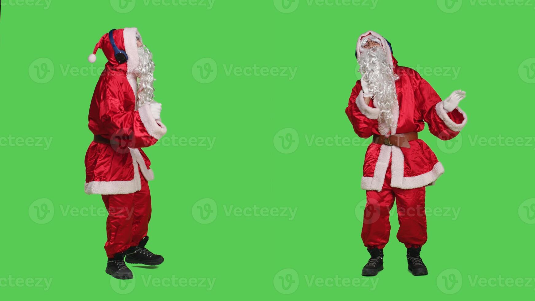 Man in red suit having fun with audio, listening to music on headphones to celebrate winter seasonal holiday. Santa with white beard dancing and enjoying songs over greenscreen backdrop. photo