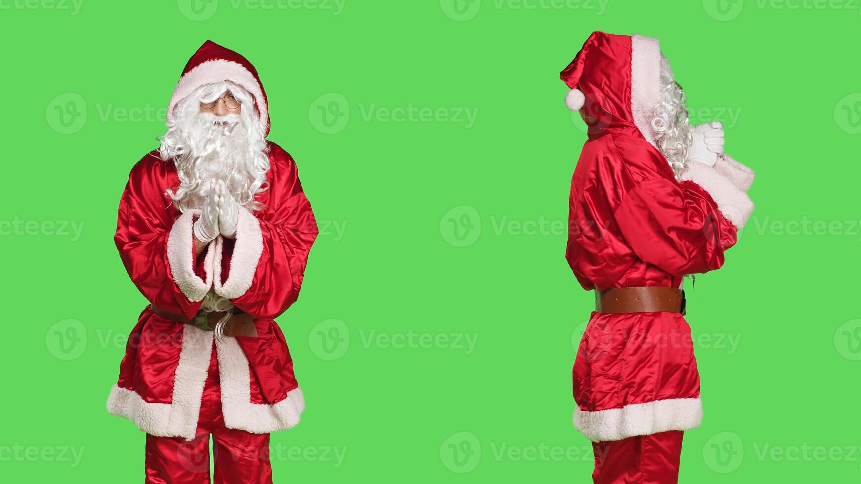 Faithful santa claus praying in studio, holding hands in a prayer and standing against greenscreen backdrop. Model portraying father christmas with festive red costume being religious. photo