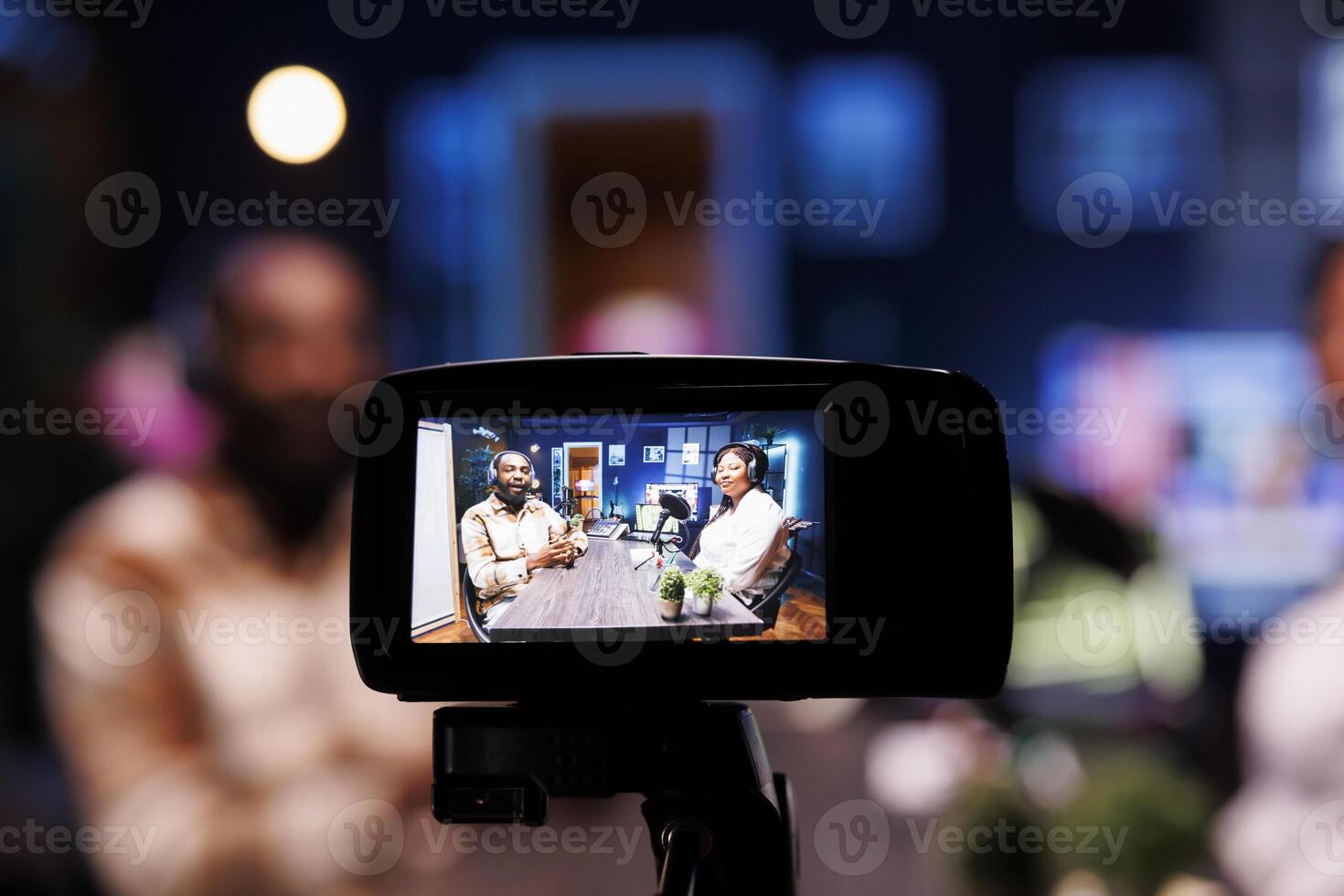 Focus on vlogging camera used by cohosts in blurry background streaming podcast during live debate on vlog channel. Close up of professional recording gear used by man and woman in studio to film show photo