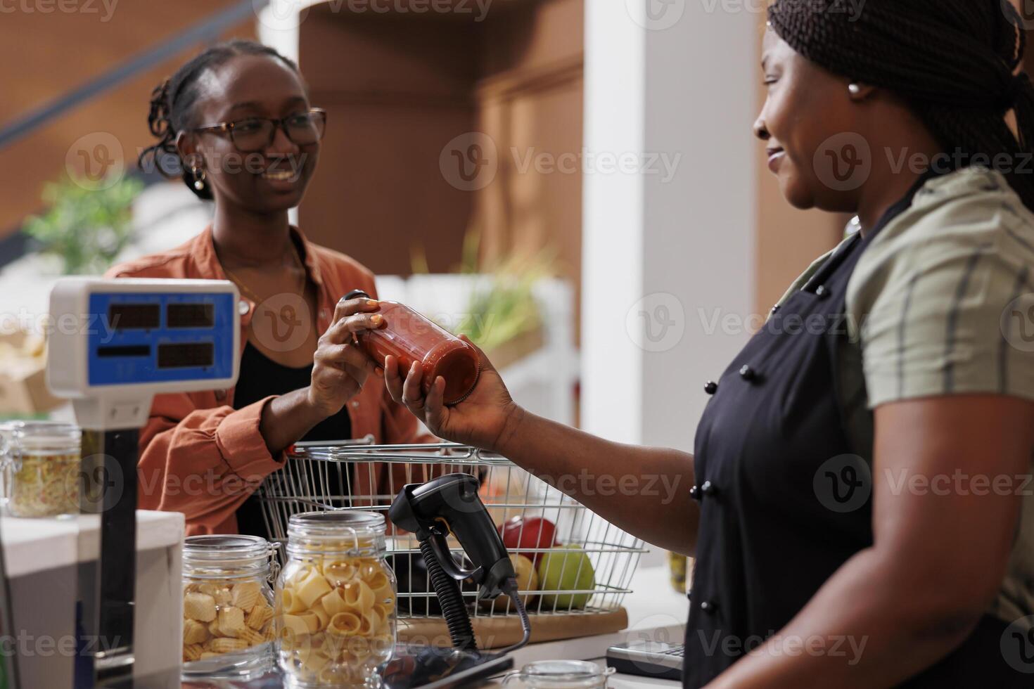 In grocery store, black woman serves a customer by selling her freshly picked organic produce. Happy customer at checkout counter gives a smiling female vendor a bottle of sauce. photo