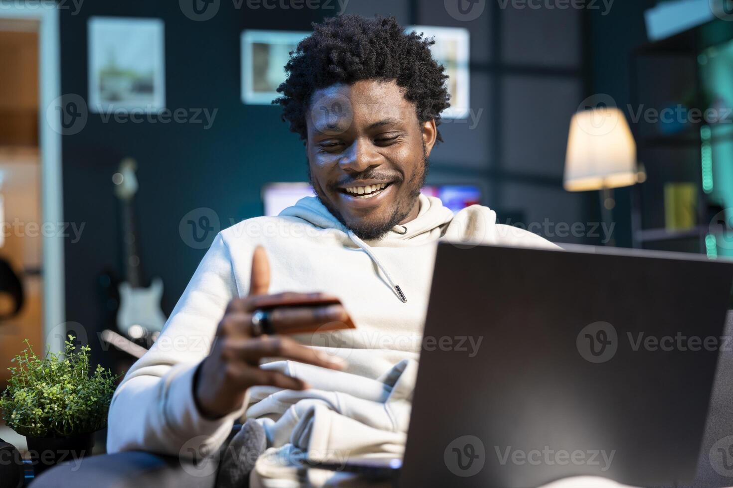 African american man laughing while adding payment method on website while in dimly lit living room. Joyous person delighted to do online shopping, looking at credit card in hand photo