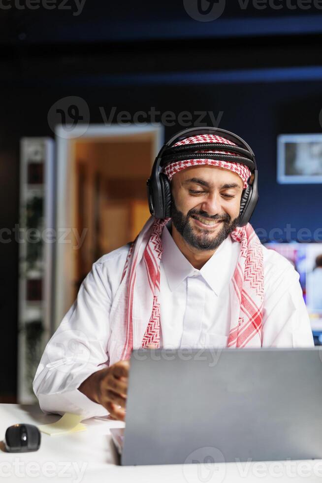 Image showcasing an Arab man with wireless headphones enthusiastically working on his personal computer. Detailed view of a cheerful Muslim student doing online research while listening to music. photo