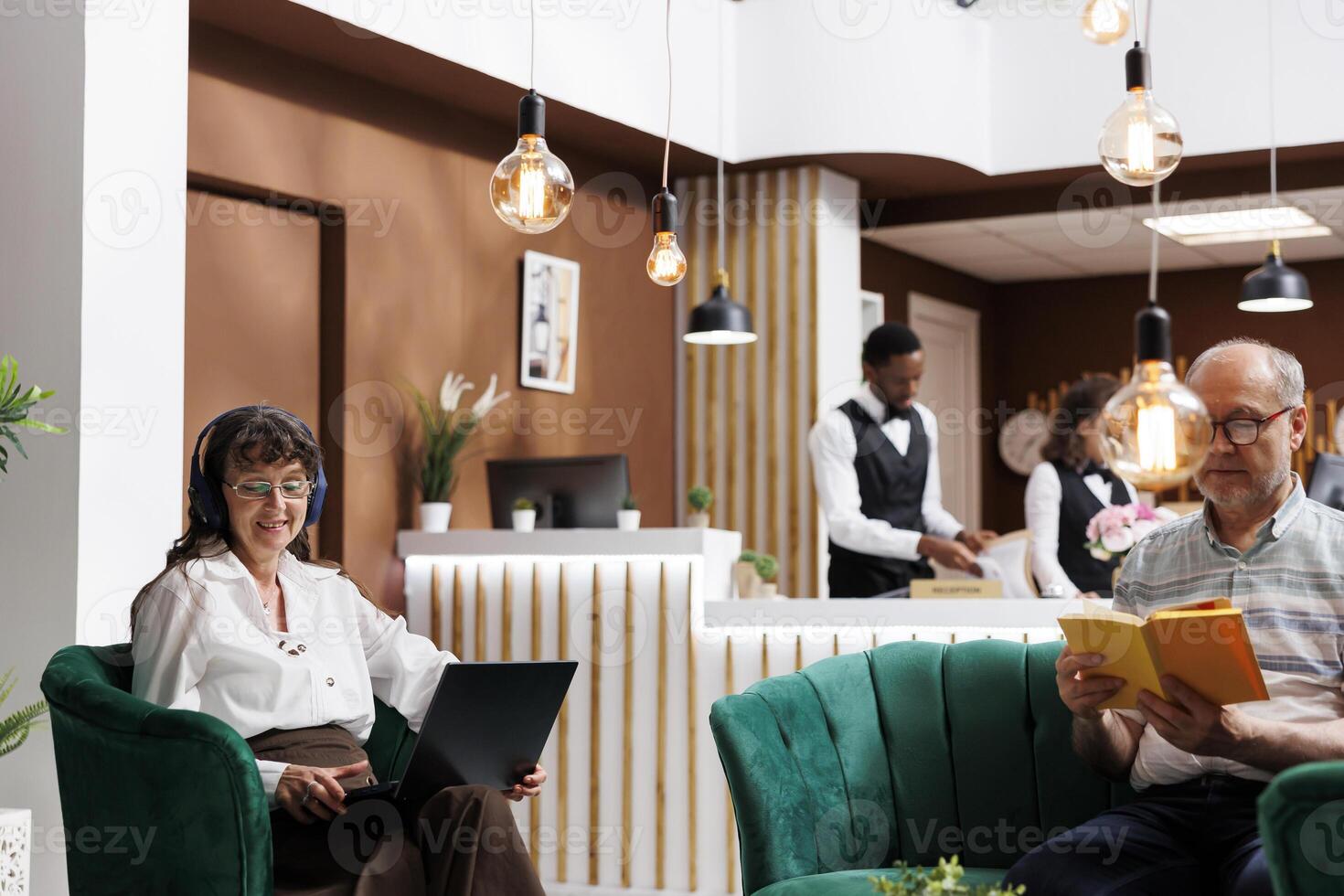 Senior woman wearing headphones sits on sofa in hotel lobby, using laptop to confirm reservation. In lounge area, male client on couch reading as retired elderly lady with headphone uses minicomputer. photo