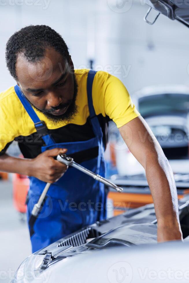 African american mechanic in car service uses torque wrench to tighten screws after replacing defective engine. Skilled garage employee uses professional tools to fix malfunctioning client automobile photo