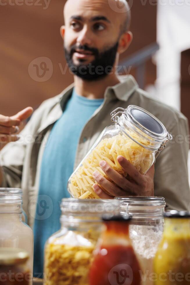 Close-up shot of middle eastern man grasping a container filled with homemade pasta in eco friendly store. Detailed image of male customer holding glass jar while browsing for other organic products. photo