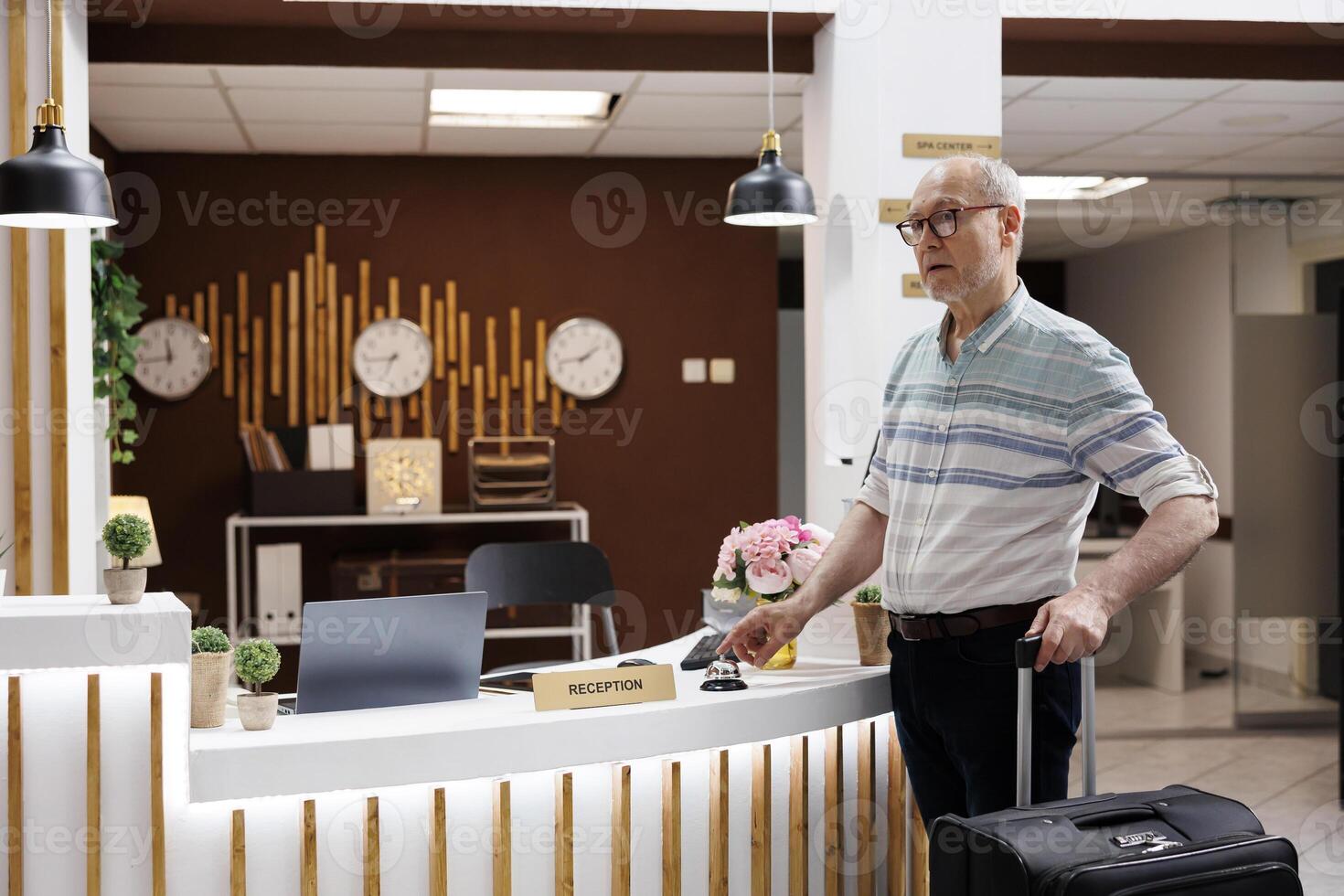 Retired old man at hotel lobby, calling for assistance by pressing concierge bell while waiting for receptionist. Elderly male tourist with suitcase lingering at front desk, ready for vacation. photo