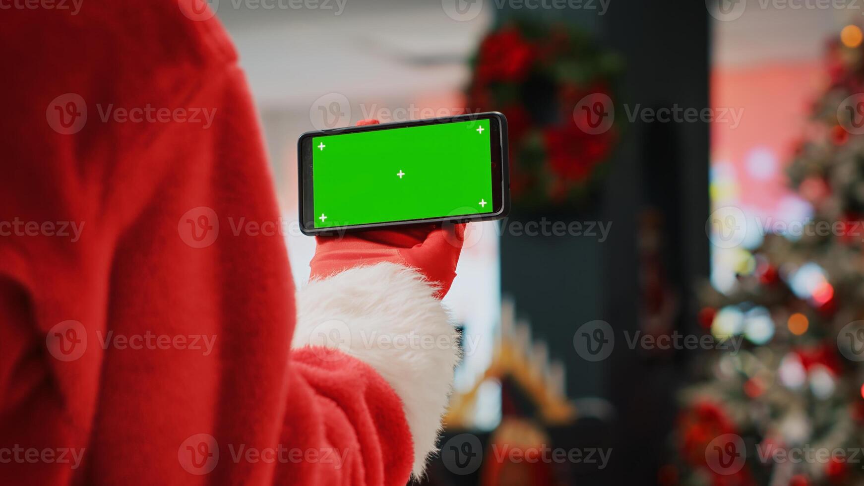 Staff assistant wearing Santa Claus suit in shopping mall adorn with Christmas decorations holding chroma key phone, taking a break from work while watching videos on green screen, close up photo