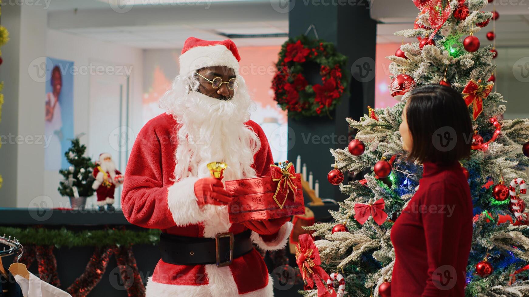 African american employee dressed as Santa Claus ringing bell and holding xmas gift next to xmas ornate tree, talking with asian client shopping in festive decorated fashion store photo
