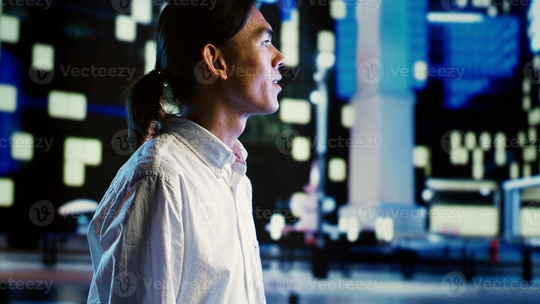 Asian man wandering around city streets at night, looking upwards to surrounding empty urban environment with no cars. Cheerful businessman taking a walk to clear his head after work photo