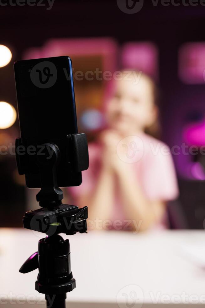 Close up of phone on tripod stabilizer used by child sponsored by brand to showcase message on children channel. Focus on smartphone recording kid in blurry background doing influencer marketing photo