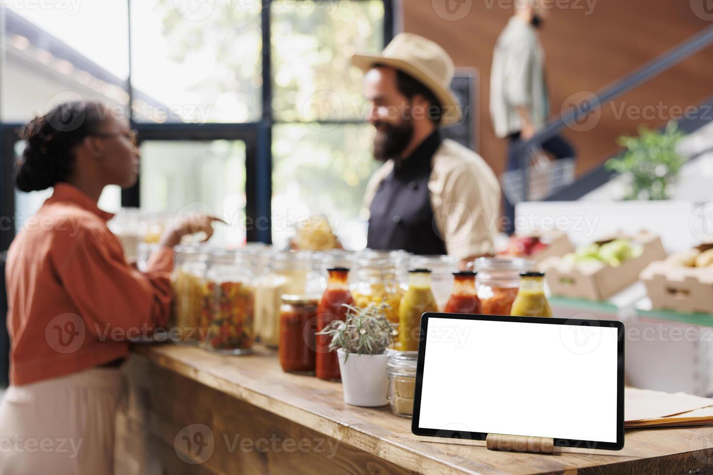 Tablet with a blank chromakey mockup template horizontally positioned near glass jars. On the counter is a smart device displaying an isolated white screen while shopkeeper converses with customer. photo