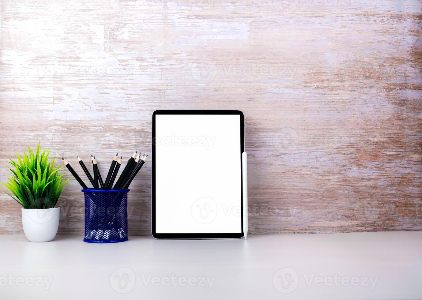 Tablet with blank screen placed on white table and leaning against wooden surface wall with office equipment. Copy space for your text. Workspace and copy space. Office desk with office supplies photo