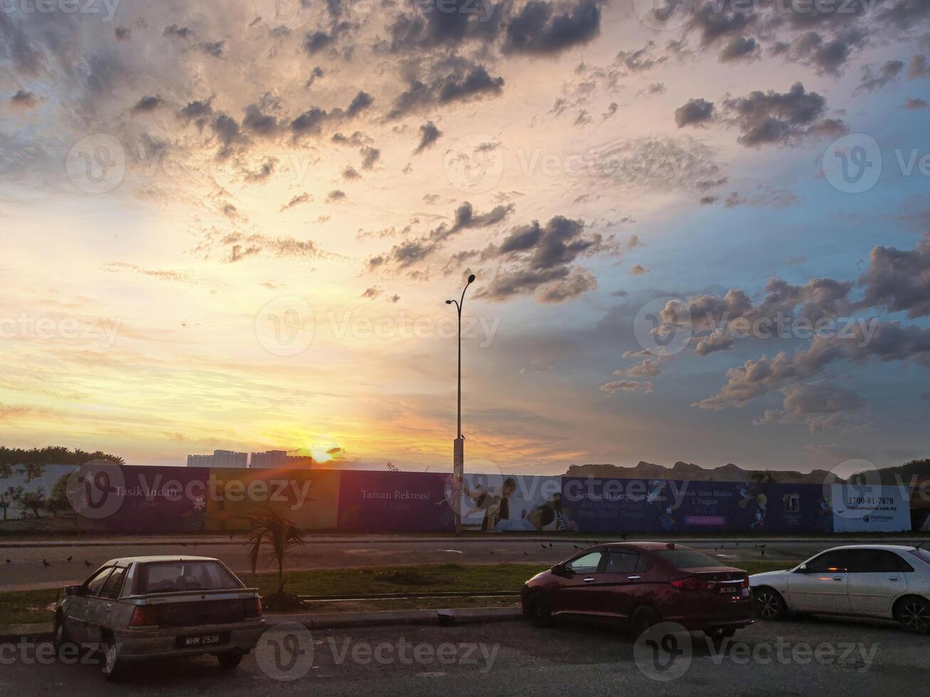The horizon glows with orange and yellow hues as the sun sets behind clouds, casting a warm light on nearby parked cars and the cityscape in Puncak Alam Malaysia. photo