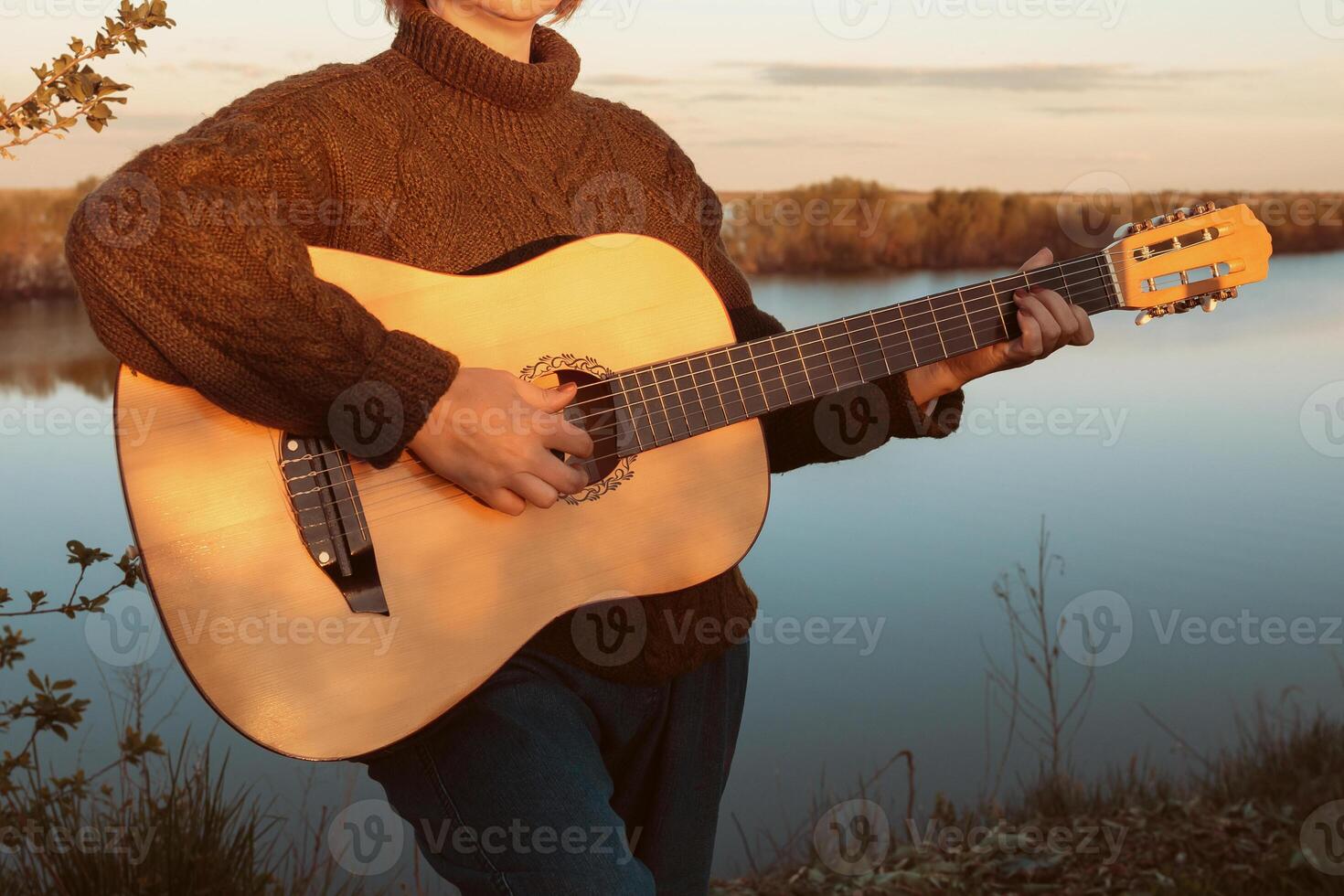 A woman in a warm sweater and pants plays a classical guitar against the background of a river at sunset photo
