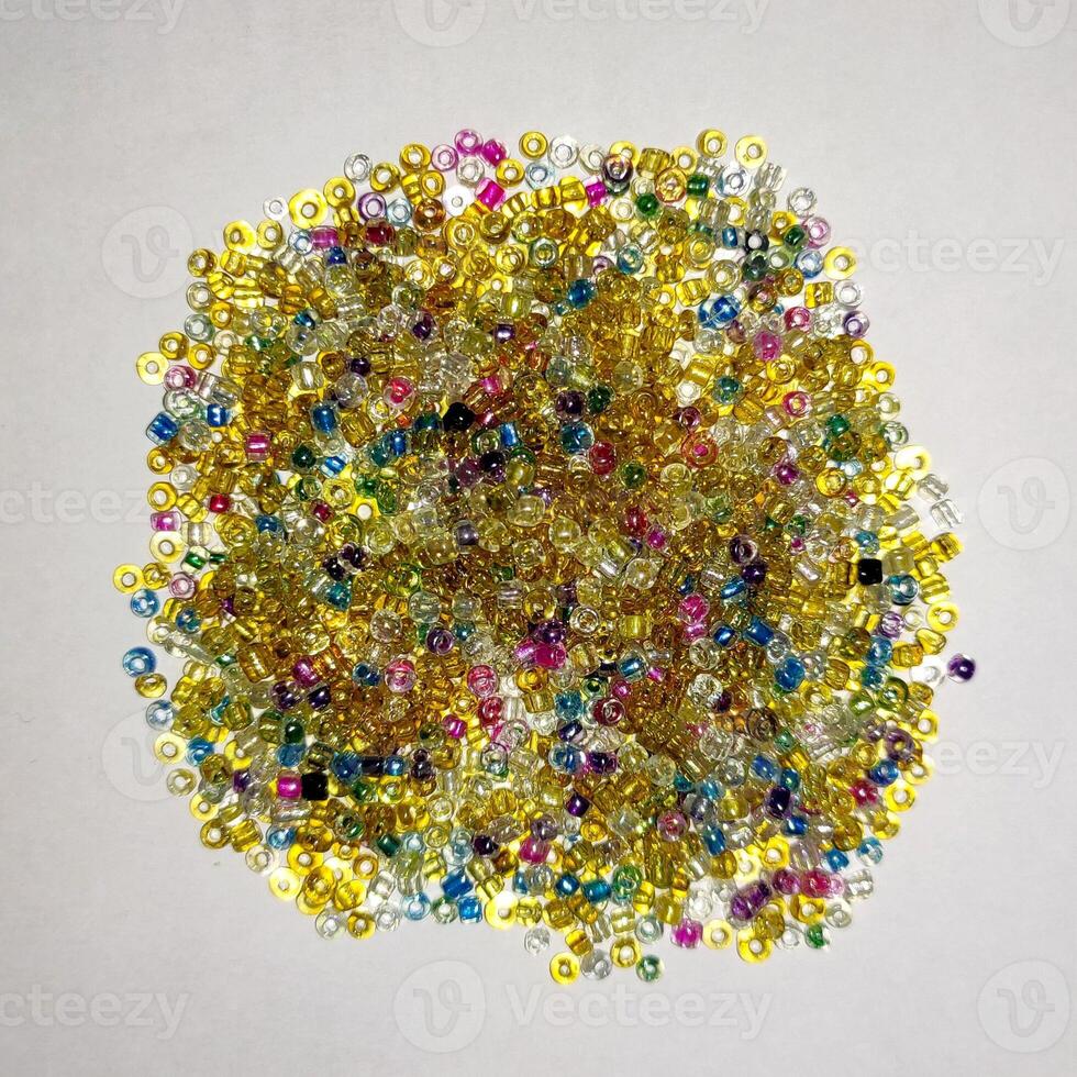 A colorful array of small, cylindrical glass beads in vibrant shades of yellow, pink, purple, green, blue, and clear is scattered across a white surface, perfect for craft projects and jewelry design. photo