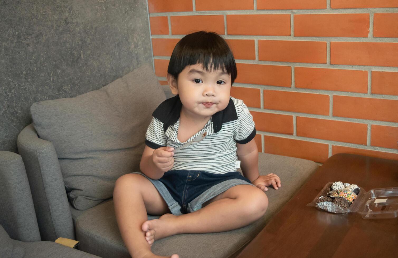 Asian cute little boy sitting on the sofa using a plasic spoon to eat mini donuts at home,enjoys eating looking at the camera and happiness photo