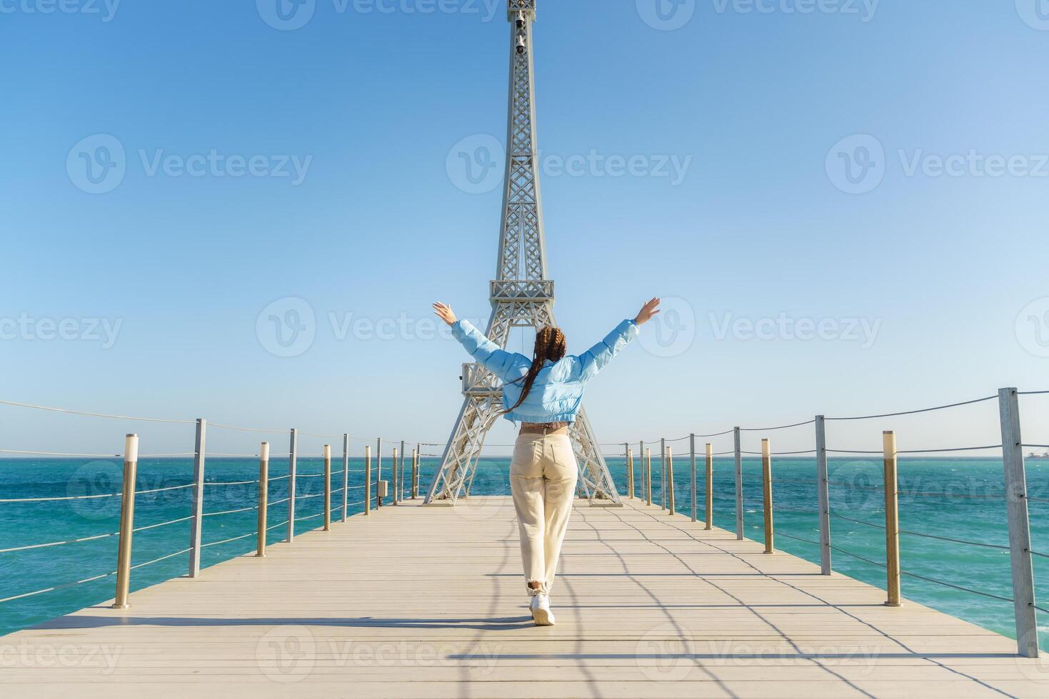 Large model of the Eiffel Tower on the beach. A woman walks along the pier towards the tower, wearing a blue jacket and white jeans. photo