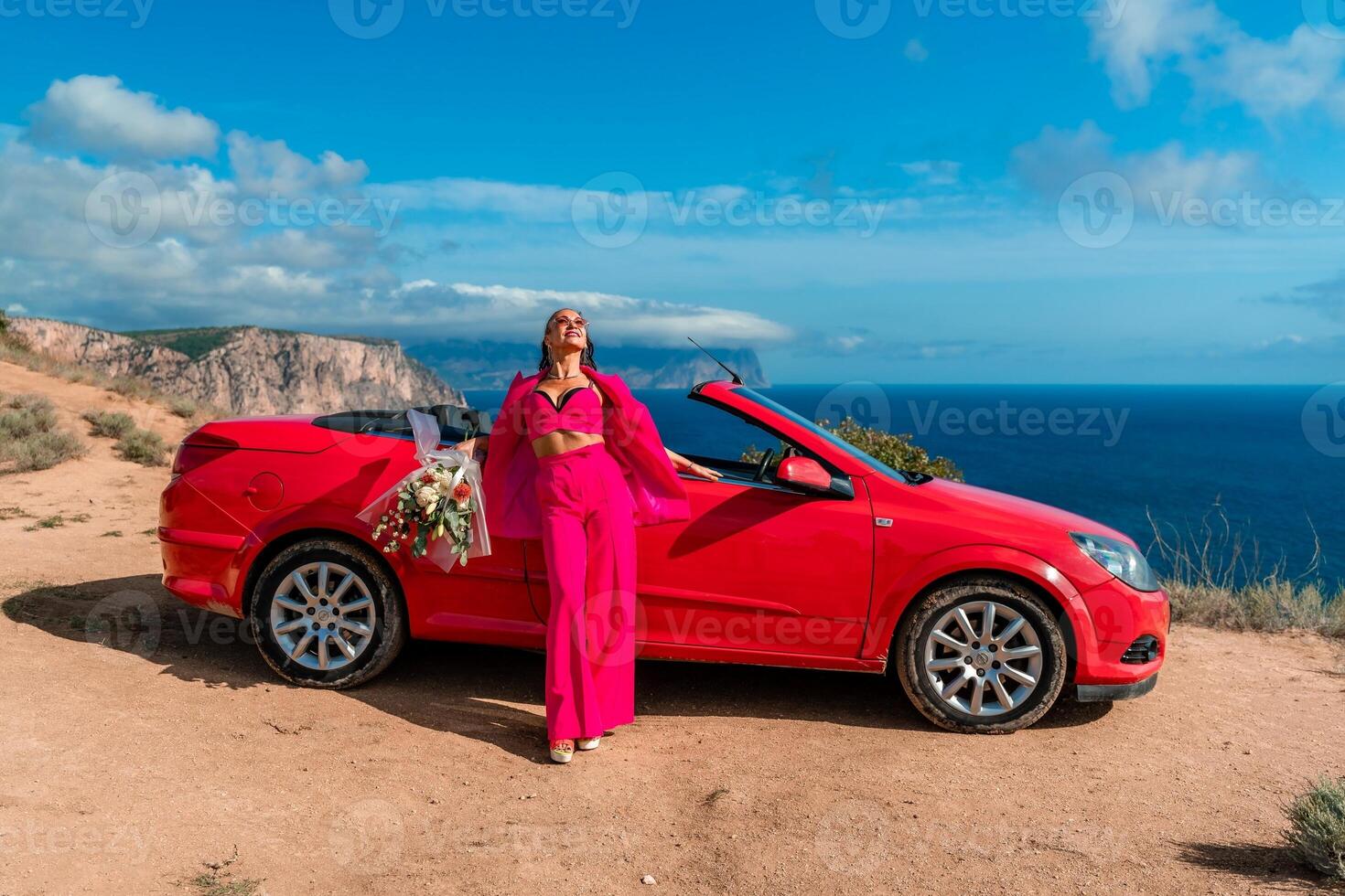 A woman stands in front of a red convertible car, holding a bouquet of flowers. Concept of freedom and adventure, as the woman is dressed in a red outfit and poses in front of the car. photo