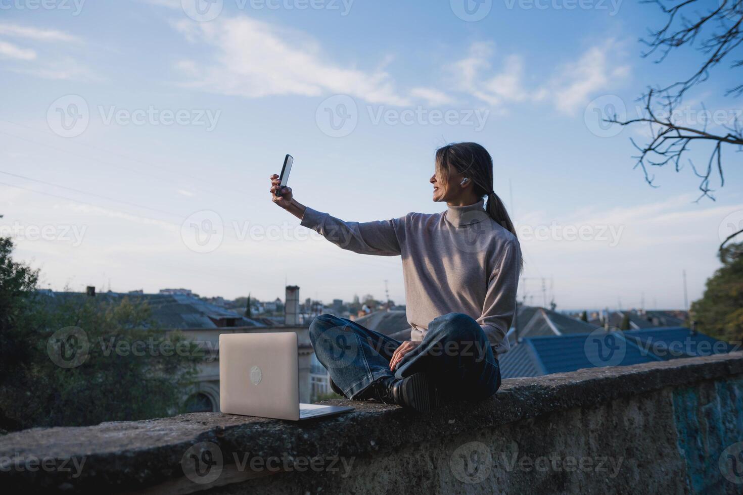 Woman freelancer uses laptop on cement wall outdoors against the sky and the roof of the city. The woman to be focused on her work or enjoying some leisure time while using her laptop. photo