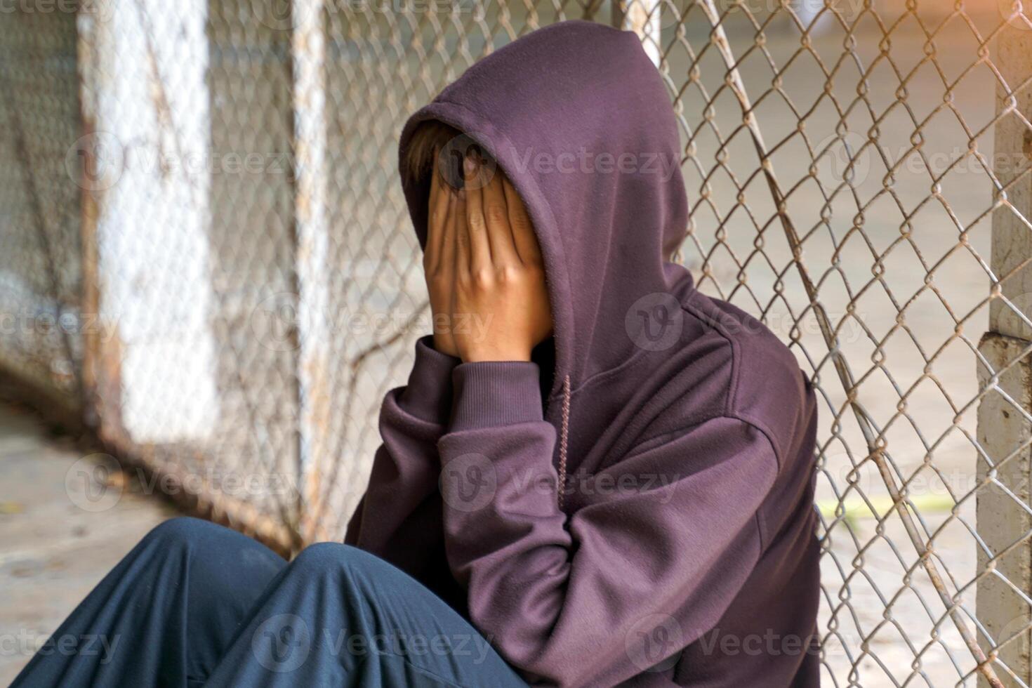 Young man wearing a hood over his head. He covered his face with his hands and sat alone against the mesh fence. photo