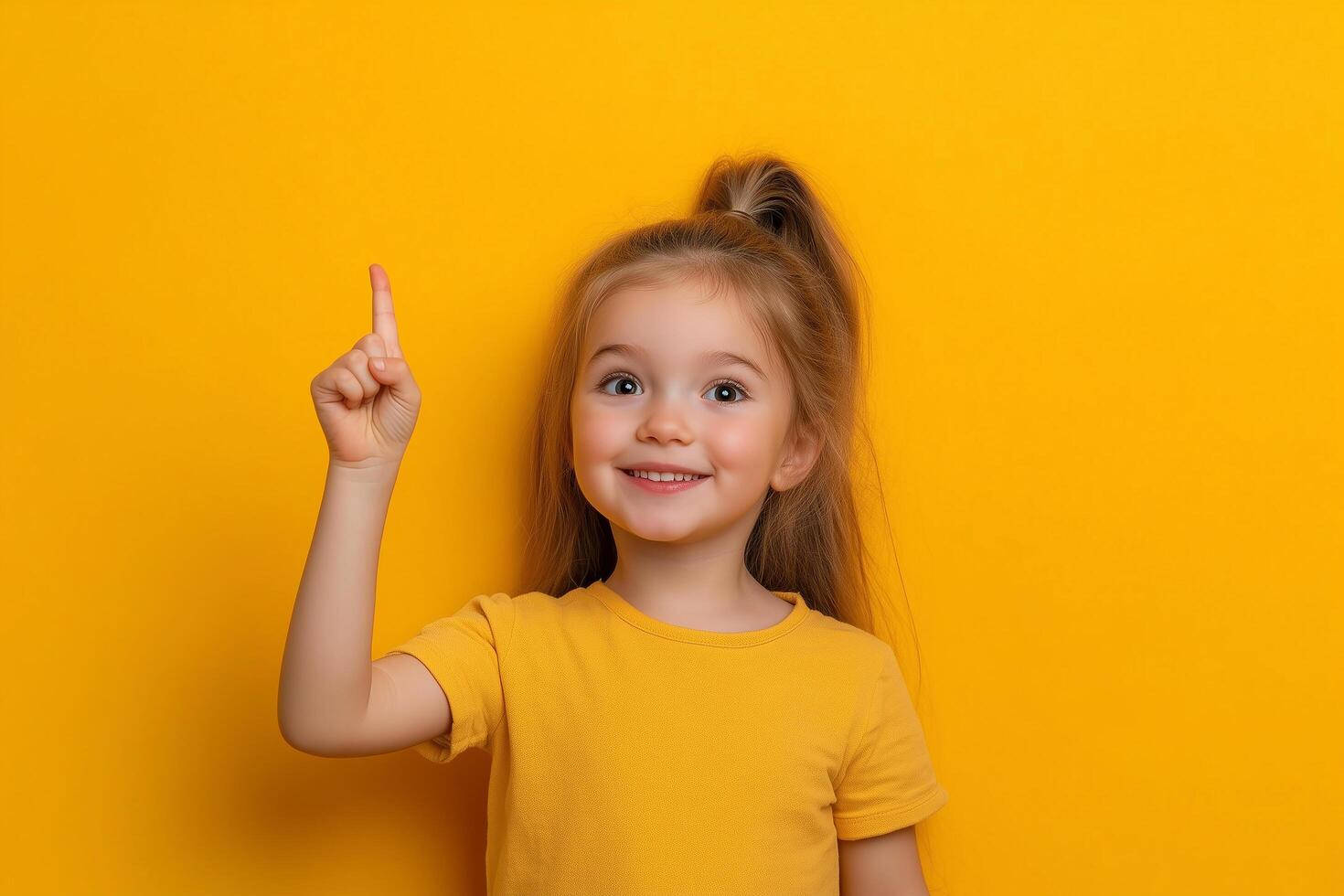 Photo of a happy young girl pointing her finger upward against a yellow background, with ample copy space