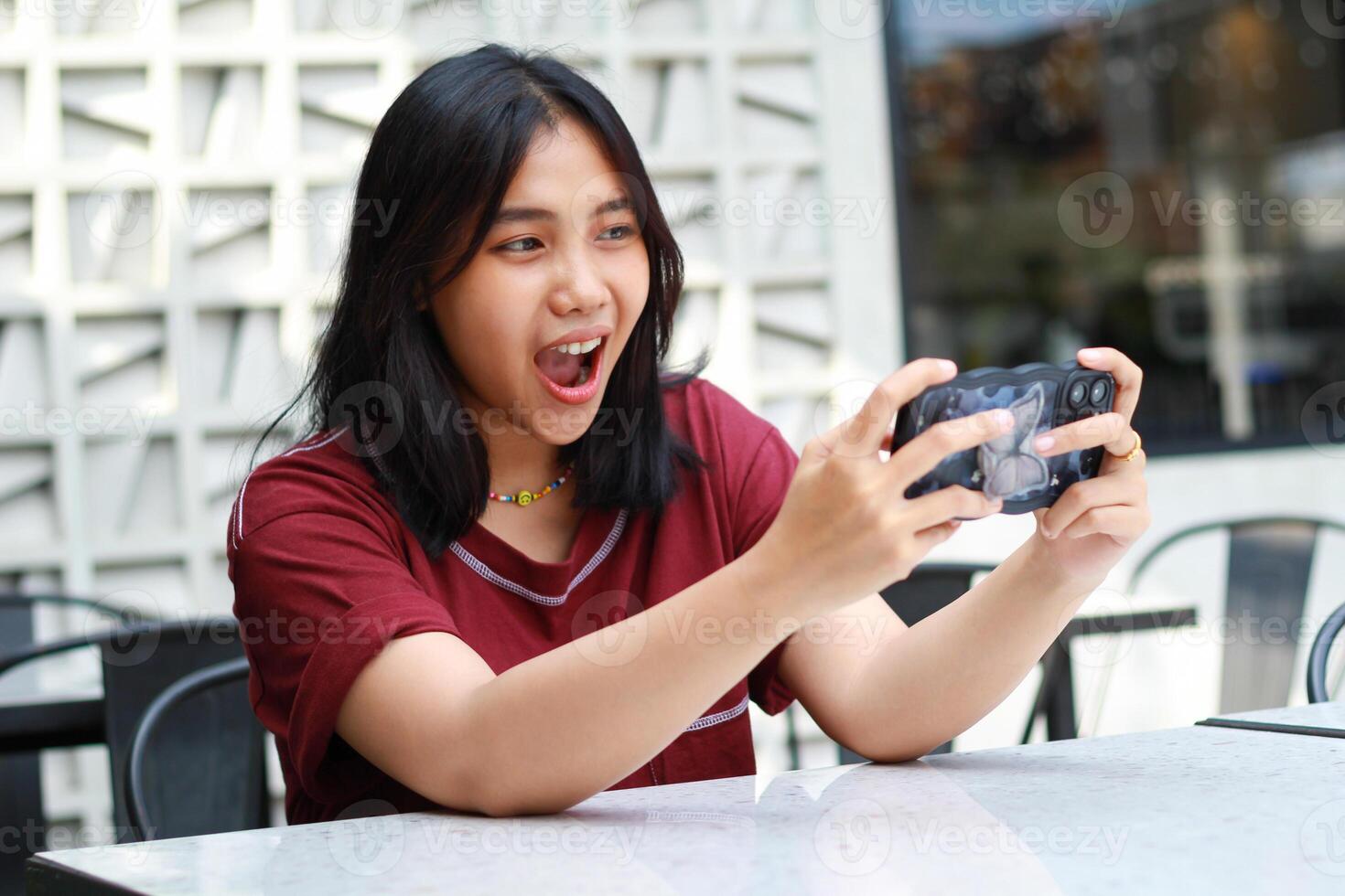 pretty asian woman excited playing game online using mobile phone sitting in outdoor cafe wearing red tshirt, shouting with open mouth photo