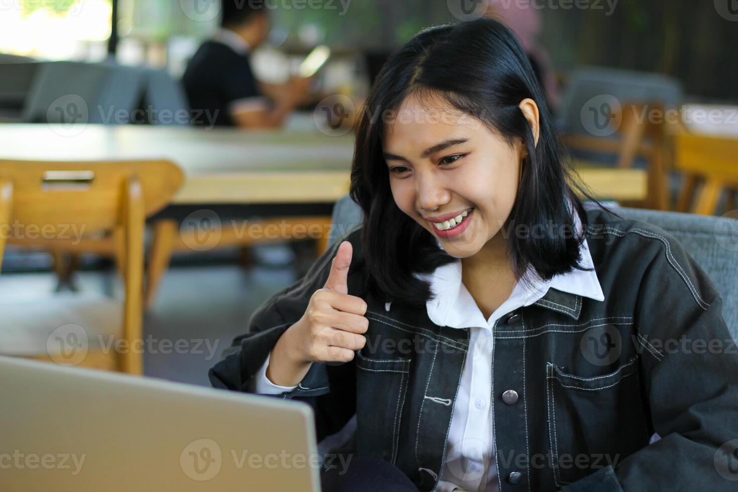 carefree asian woman sitting in cafe showing thumb up and laughing while doing virtual online meeting with client in cafe photo