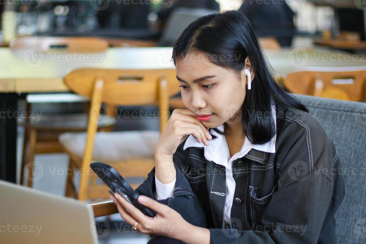 pretty and charming asian woman feeling bored scrolling social media using smartphone in cafe with laptop over desk photo