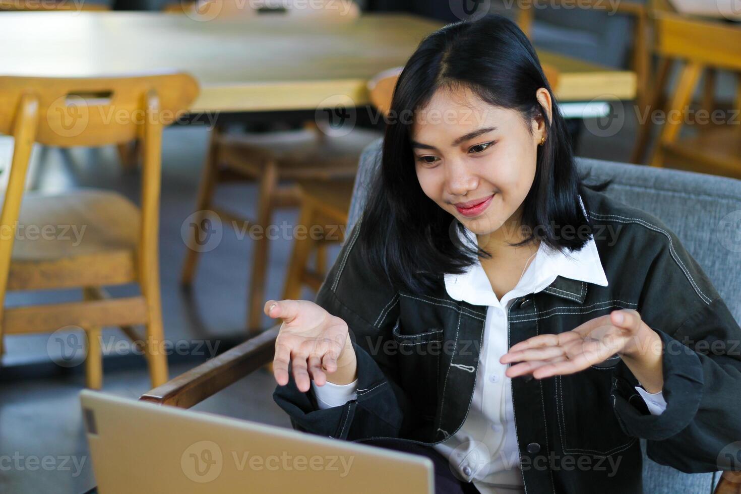 pretty asian woman looks happy doing virtual presentation with client in cafe using laptop, raising palms showing presenting gesture photo