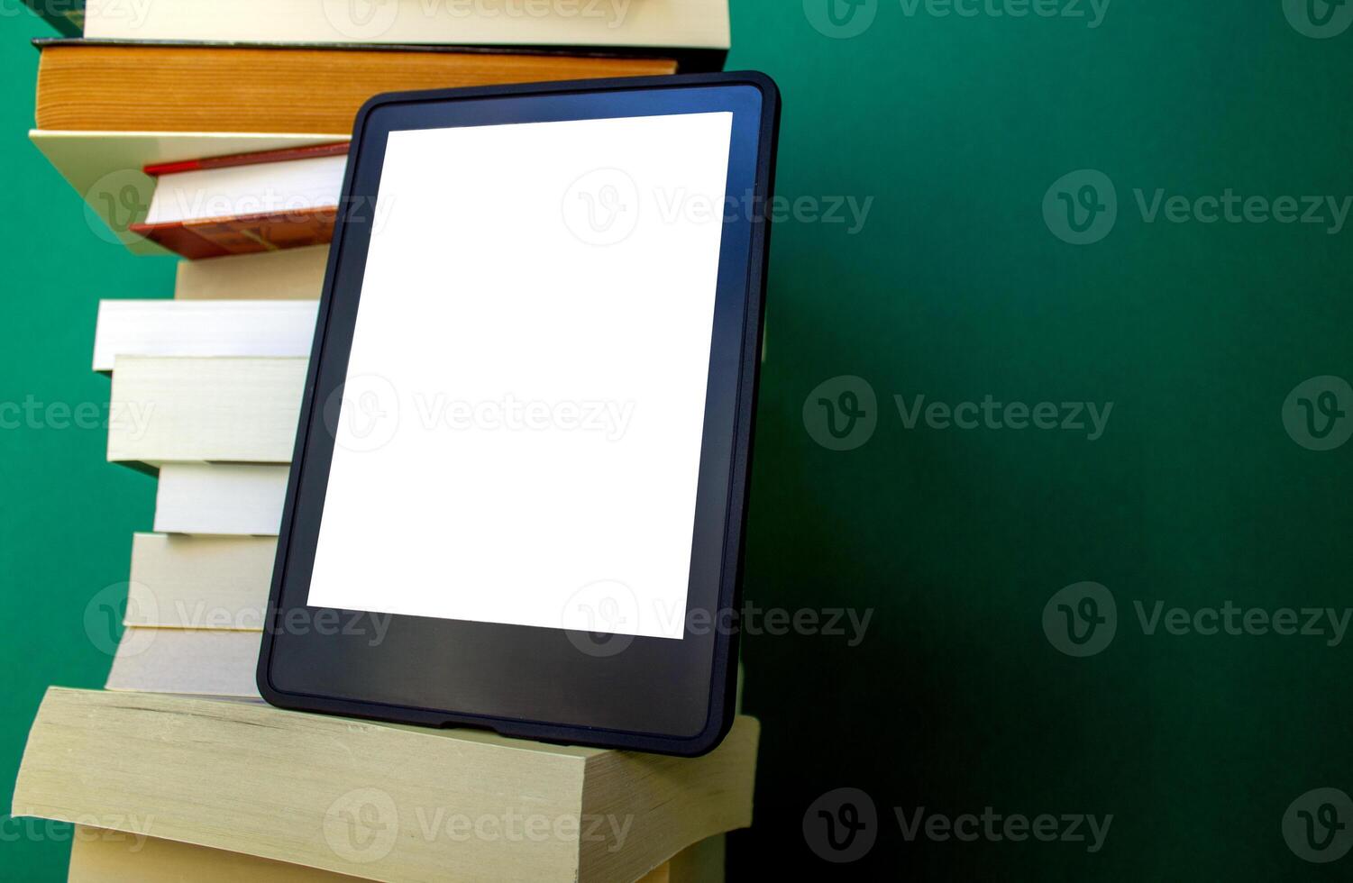 an electronic book next to a stack of paper books on a dark green background. one e-book replaces many regular books photo