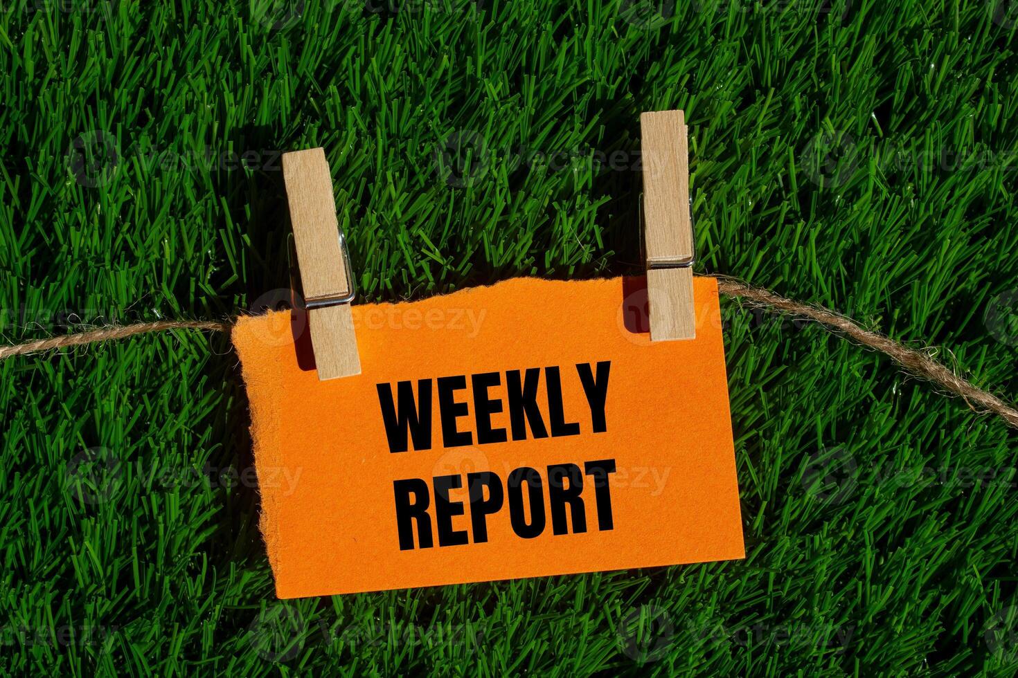 Weekly report message written on ripped torn orange paper piece with green grass background. Conceptual weekly report symbol. Copy space. photo