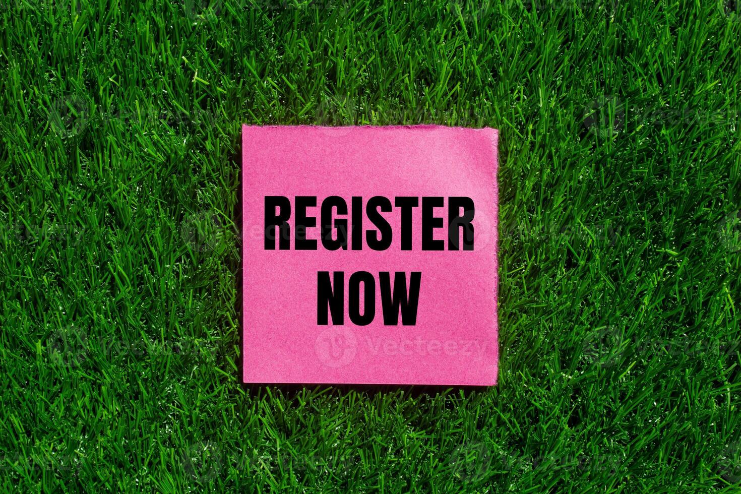 Register now message written on ripped torn pink paper with green grass background. Conceptual register now symbol. Copy space. photo