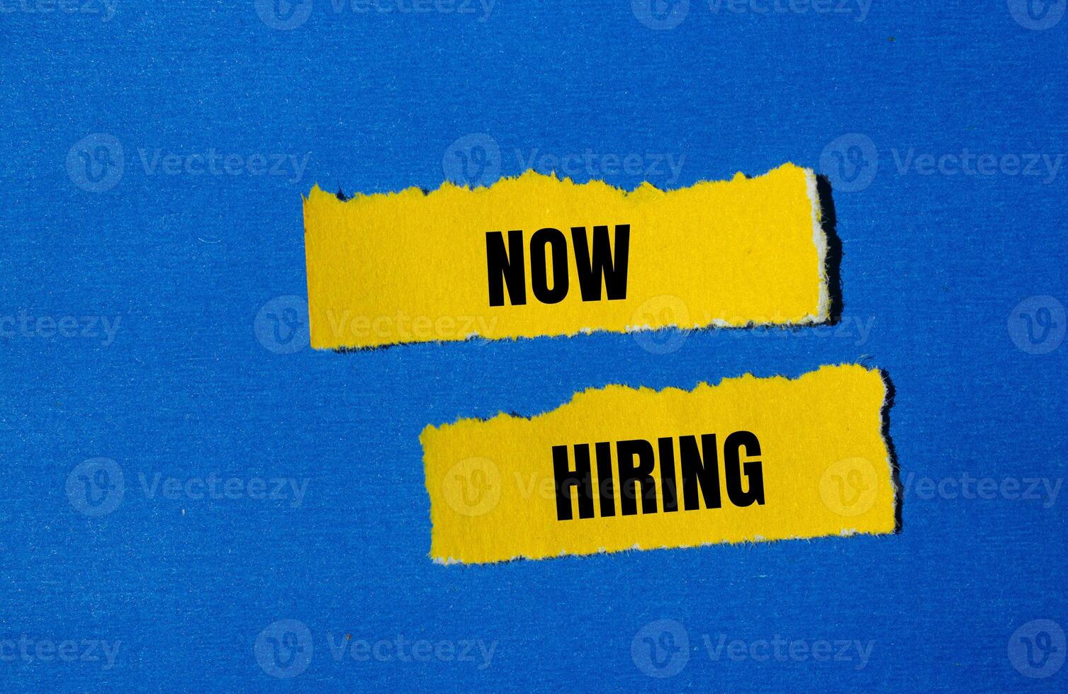 Now hiring message written on ripped yellow torn paper pieces with blue background. Conceptual now hiring symbol. Copy space. photo