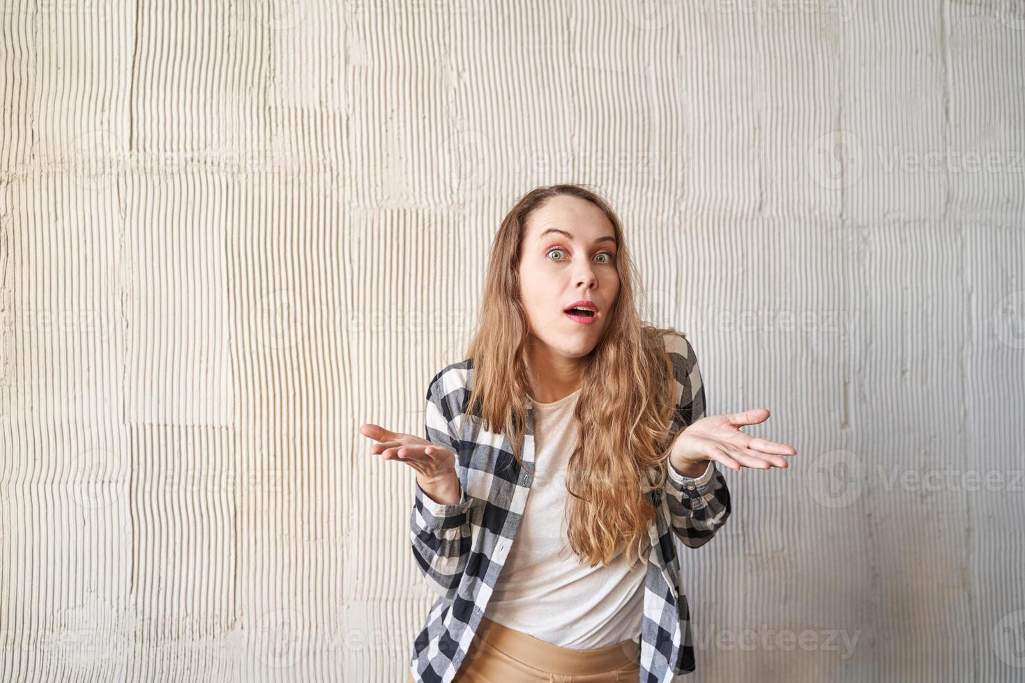 Confused woman shrugging against textured wall - expressions of uncertainty and curiosity in casual checkered shirt scene. photo