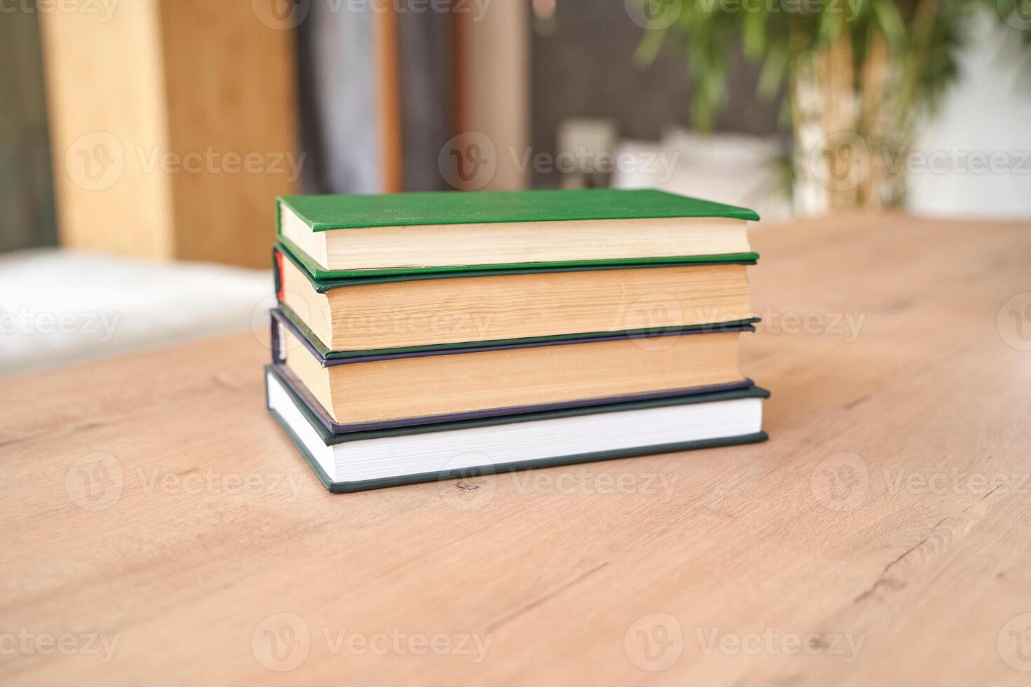 Stack of hardcover books on wooden table in cozy indoor setting with greenery and natural lighting. photo