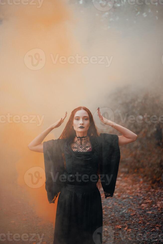 Goth woman, red-haired witch in autumn forest in fog in black dress, Halloween. Mystical atmosphere photo