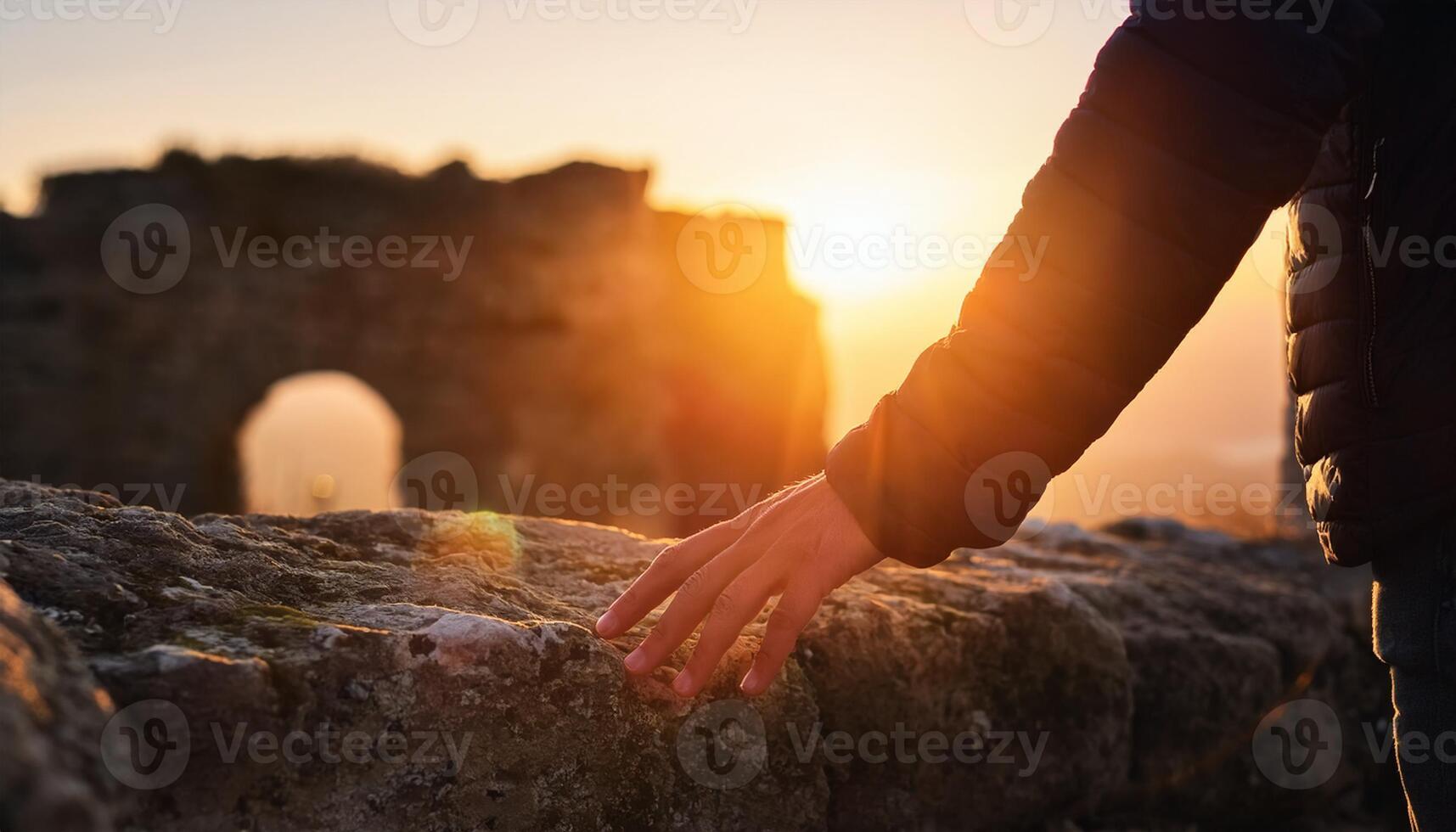 A person's hand gently touches an ancient stone wall, with warm sunlight and historic ruins in the background during sunset. photo