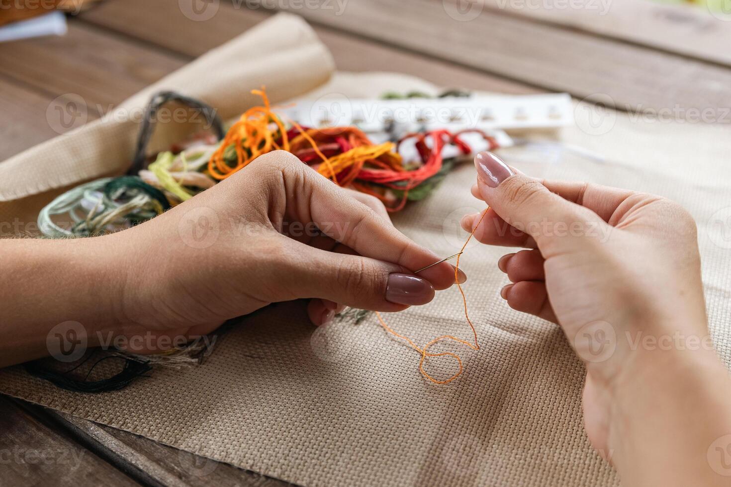 Putting the thread through the eye of the needle. Close-up of the cross stitching process. In the background is a wooden table with a stack of multi-colored embroidery threads and embroidery scissors. photo