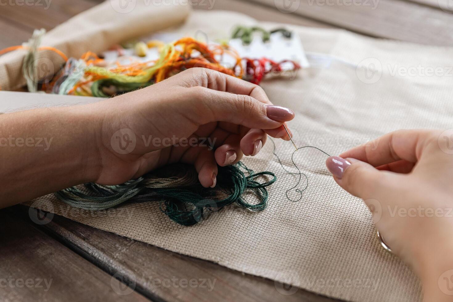 Putting the thread through the eye of the needle. Close-up of the cross stitching process. In the background is a wooden table with a stack of multi-colored embroidery threads and embroidery scissors. photo