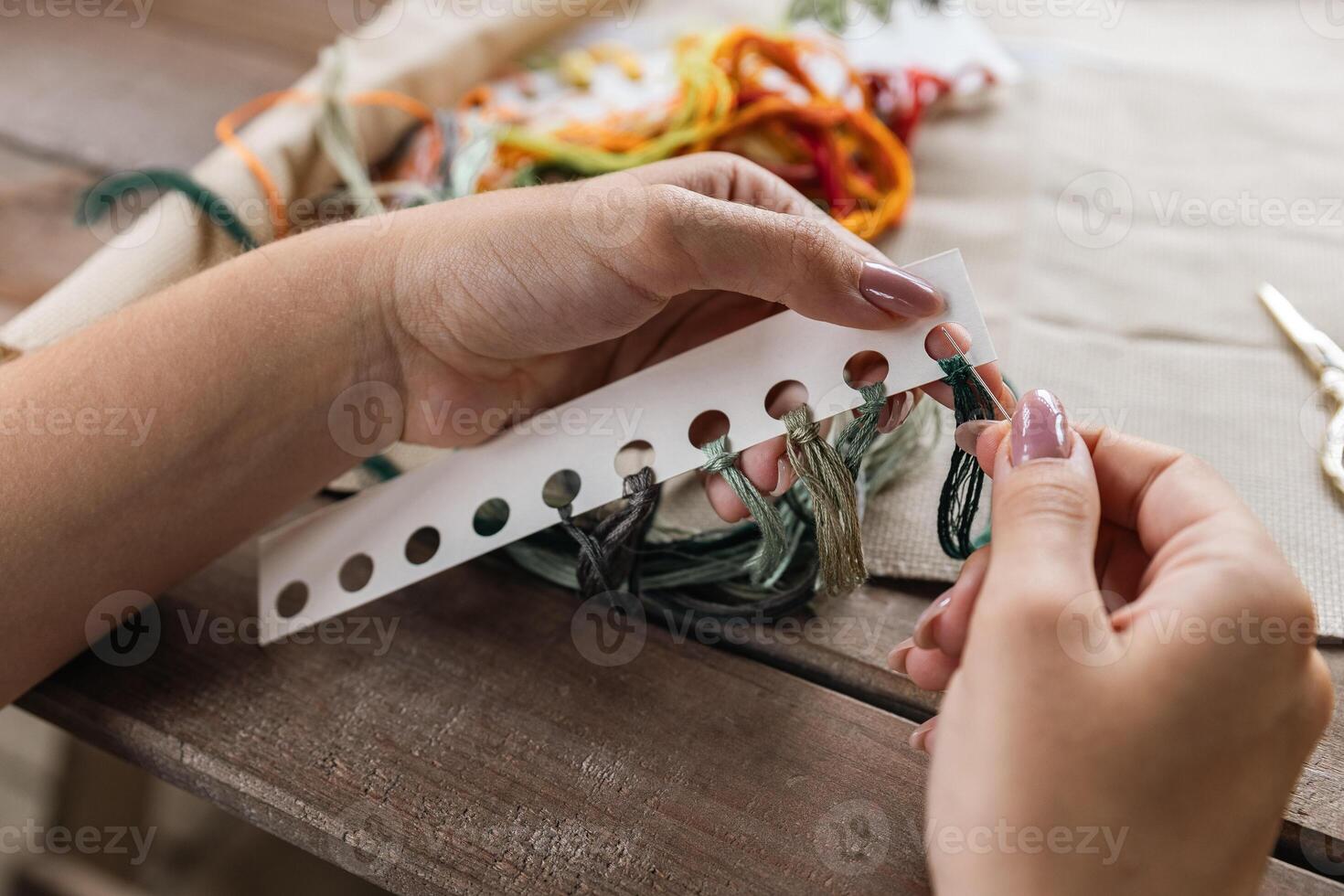 Putting the thread through the eye of the needle. Close-up of the cross stitching process. In the background is a wooden table with a stack of multi-colored embroidery threads and embroidery scissors. photo
