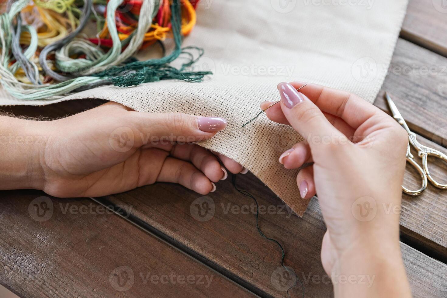 A woman doing cross stitching. Close-up of hands with a needle threading the thread through the fabric. Embroidery of a picture. In the background are colorful embroidery threads. photo