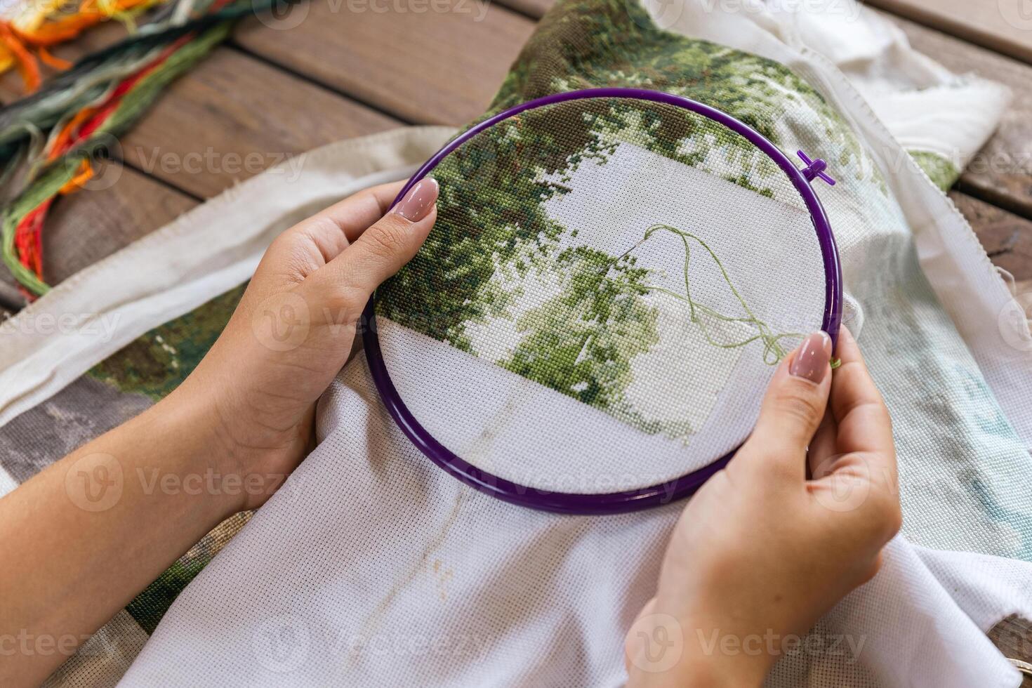 A girl cross-stitching. Close-up of hands carefully maneuvering a needle through the fabric. A purple embroidery hoop holds the fabric. Close-up of hands with a needle threading the fabric. photo