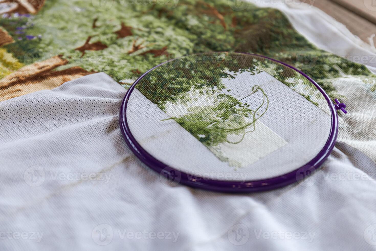 Close-up of a cross-stitch embroidery project in progress. The needle is threaded with green floss and is partially inserted in the fabric. The hoop is purple, and the fabric is white. photo