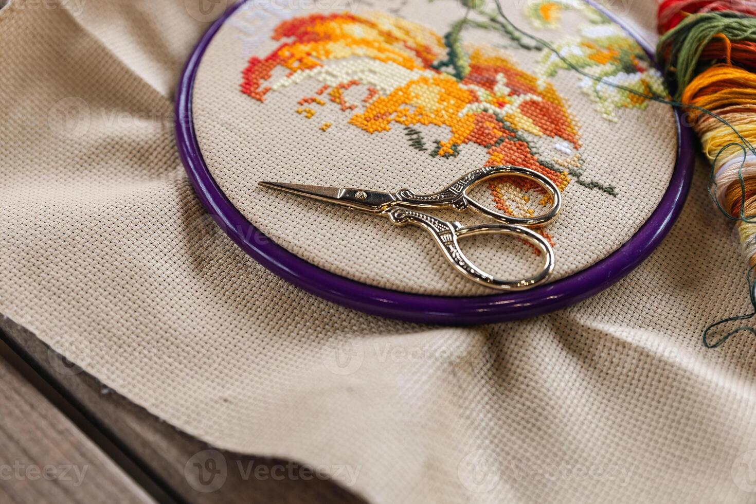 Cross-stitch, with an embroidery hoop containing a partially completed design of an orange and white flower. A pair of gold embroidery scissors rests on top of the hoop. photo