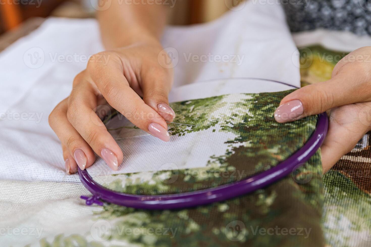 A girl cross-stitching. Close-up of hands carefully maneuvering a needle through the fabric. A purple embroidery hoop holds the fabric. Close-up of hands with a needle threading the fabric. photo