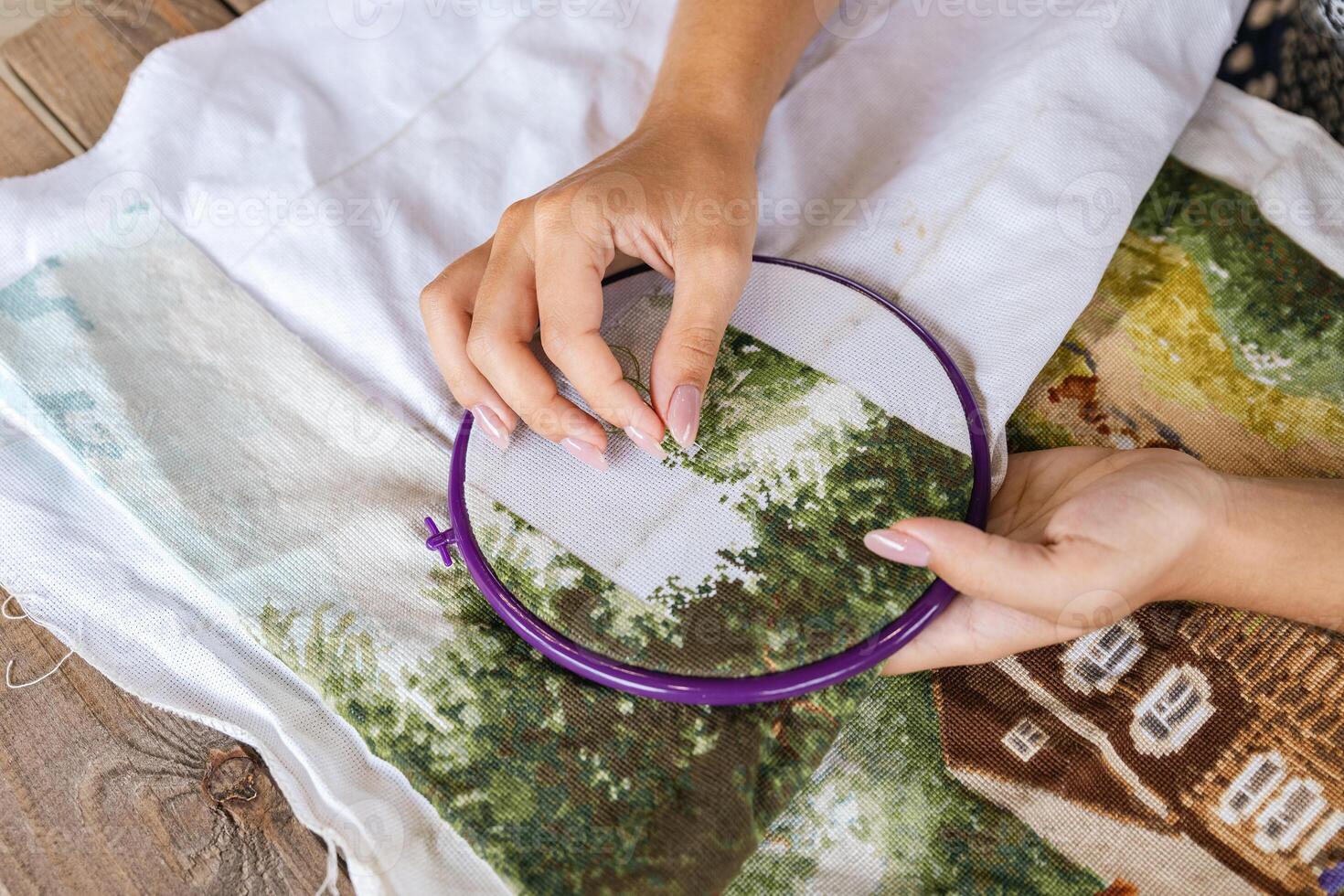 A girl cross-stitching. Close-up of hands carefully maneuvering a needle through the fabric. A purple embroidery hoop holds the fabric. Close-up of hands with a needle threading the fabric. photo