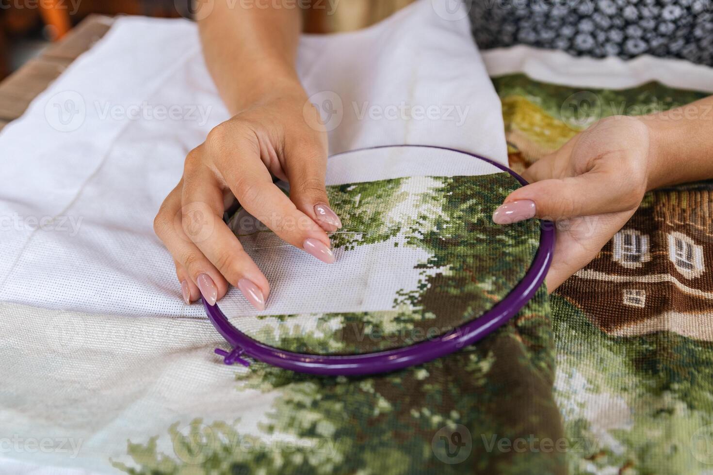 A girl cross-stitching. Close-up of hands carefully maneuvering a needle through the fabric. A purple embroidery hoop holds the fabric. Close-up of hands with a needle threading the fabric. photo